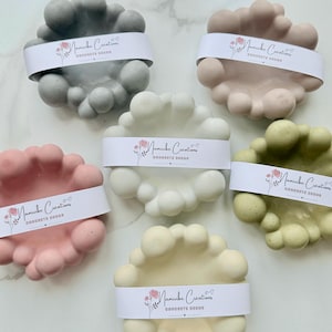 Op de afbeelding: Een verzameling wolk-vormige betonnen decoratiestukken in verschillende kleuren: grijs, roze, wit, geel en groen. Elk stuk is omwikkeld met een witte band met de tekst "Maminbe Creations CONCRETE DECOR".