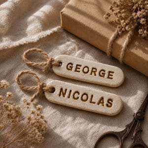 Può includere: Due etichette rettangolari in ceramica color bianco sporco con bordi arrotondati, ciascuna dotata di un occhiello in corda di juta. L'etichetta superiore reca il nome GEORGE e quella inferiore il nome NICOLAS in lettere maiuscole scure incise.