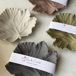 Puede incluir: Tres platos de hoja de hormigón en diferentes colores: beige, verde y gris. Cada plato tiene una etiqueta blanca con el texto "Namuibe Creations Concrete Decor" y un pequeño diseño floral.