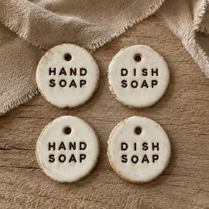 May include: Four circular, off-white ceramic tags with a hole at the top. Two tags are stamped with the black text HAND SOAP, and two tags are stamped with the black text DISH SOAP. The ceramic pieces have a slightly uneven, hand-crafted edge.