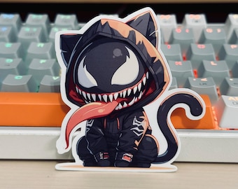 May include: A sticker depicting a cartoon cat character in a black hoodie, with a Venom-inspired design. The cat has a large, toothy grin, a long red tongue, and white eyes. The sticker is placed in front of a keyboard.