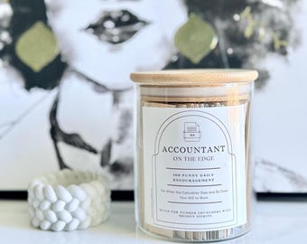 May include: A glass jar with a wooden lid, labeled "ACCOUNTANT ON THE EDGE" containing small cards for daily encouragement. A white, braided decorative object sits to the left of the jar. The background features a black and white image.