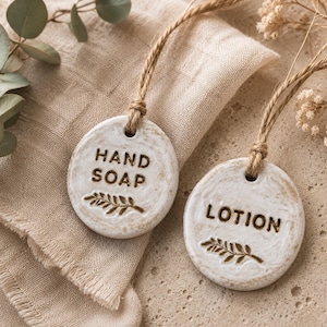 Op de afbeelding: Twee ronde, gebroken witte keramische labels met een verweerde, textuurrijke afwerking. Elk label heeft bruine, in reliëf aangebrachte tekst met HAND SOAP en LOTION, een kleine takafbeelding onder de woorden en een lus van jute touw.