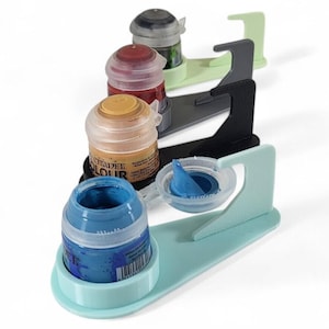 Könnte beinhalten: Vier Farbtöpfe in verschiedenen Farben werden auf einem Satz von vier Farbtöpfchenhaltern gezeigt. Die Halter sind aus hellblauem, dunkelgrauem, hellgrünem und schwarzem Kunststoff. Die Farbtöpfe sind mit "Citadel Colour" beschriftet.