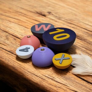 Könnte beinhalten: Ein Satz von sechs bunten, runden Klettergriffen mit verschiedenen Zahlen und Buchstaben. Die Griffe sind aus Kunststoff und haben eine strukturierte Oberfläche. Die Griffe sind auf einer Holzoberfläche zu einem Stapel angeordnet.