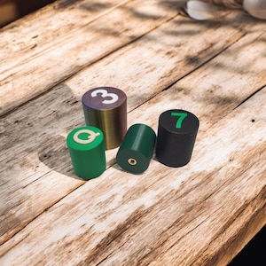 Könnte beinhalten: Vier runde, zylindrische Spielsteine mit Zahlen und Buchstaben auf der Oberseite. Die Steine sind grün, schwarz und gold. Die Zahlen sind 3, 7 und Q.