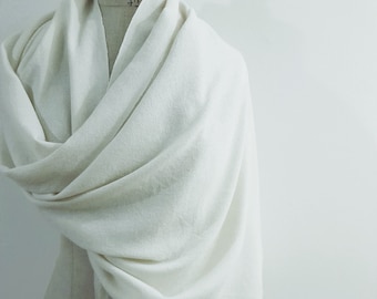 Puede incluir: Una bufanda color crema drapeada sobre un maniquí. La bufanda está hecha de una tela suave y tejida y presenta flecos en el borde inferior. La bufanda está envuelta alrededor del cuello y los hombros del maniquí, creando un aspecto fluido y elegante.