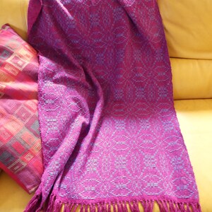 Op de afbeelding: Een magenta geweven sprei met een bloemmotief en franjes. De deken is over een gele bank gedrapeerd, met een kussen met patroon links. De sprei heeft een zachte textuur en een decoratief ontwerp.
