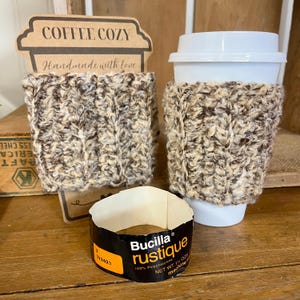May include: Two coffee cozies made of a brown and beige knit fabric. One cozy is on a white coffee cup with a lid. The other cozy is on a cardboard sign that reads "COFFEE COZY Handmade with love". A Bucilla label is visible.