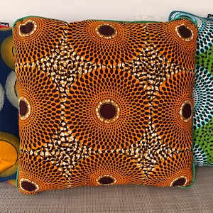May include: Square cushion with an orange and brown pattern featuring a repeating circular design. The cushion has a green trim and is part of a set with other patterned cushions.
