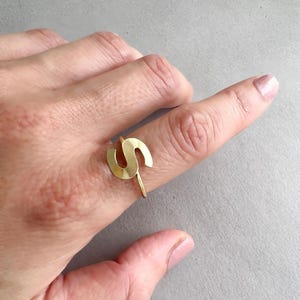 May include: A gold-colored ring in the shape of the letter "S" is displayed on a finger. The ring is made of a thin metal and has a minimalist design. The background is a neutral gray.