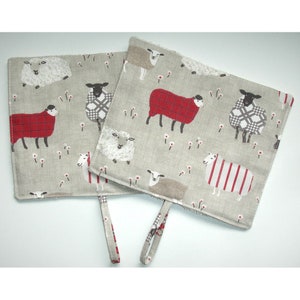 May include: Two square pot holders with a sheep pattern. The fabric is a light grey with various sheep designs in red, black, white, and grey. Each pot holder has a small loop for hanging.
