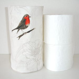 May include: A white fabric cover featuring a printed robin bird with a red breast perched on a branch and light gray floral line art, positioned next to two stacked white toilet paper rolls with a subtle embossed texture on the paper surface.