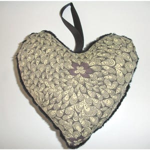 May include: Heart-shaped decorative pillow with a black fur trim and a black ribbon loop. The pillow features a cream-colored pattern with dark brown details and a purple floral accent in the center. A decorative item.