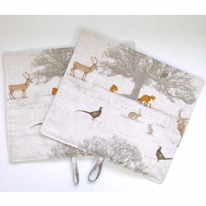 May include: Two beige linen kitchen pot holders with a woodland scene design. The design features a deer, a fox, a pheasant, a squirrel, and a rabbit. The pot holders have a loop handle.