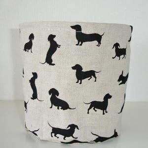 May include: A cylindrical storage basket made of light beige fabric featuring a repeating pattern of black dachshund silhouettes in various poses, including standing, sitting, and walking, distributed across the entire exterior surface.