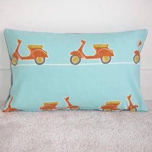 May include: Rectangular throw pillow with a light blue background and a repeating pattern of orange scooters with yellow and gray accents. The pillow is resting on a light gray surface.