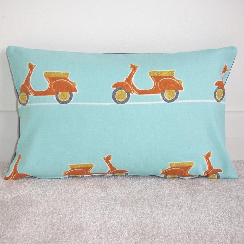 May include: Rectangular throw pillow with a light blue background and a repeating pattern of orange scooters with yellow and gray accents. The pillow is resting on a light gray surface.