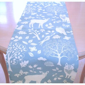 May include: A blue table runner with a white woodland pattern. The pattern includes trees, deer, birds, foxes, owls, mushrooms, and leaves.