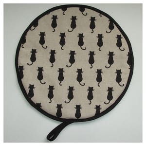 May include: Round, beige pot holder with a black trim and a black cat pattern. The pot holder has a black loop handle.