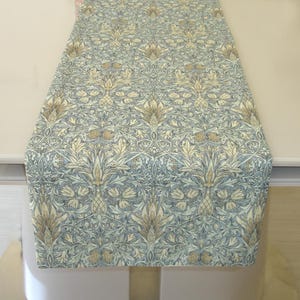May include: A rectangular table runner featuring a repeating botanical pattern in shades of muted blue, cream, and light brown. The design consists of intricate floral motifs, stylised leaves, and interconnected vines covering the entire fabric surface.