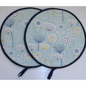 May include: Two round, floral-patterned pot holders. The pot holders have a light blue background with white, navy, and yellow floral designs. Each pot holder has a navy blue trim and a navy blue loop for hanging.