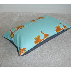 May include: A rectangular throw pillow with a light blue background and a repeating pattern of orange scooters. The pillow has a dark gray border and is designed for decorative use. The scooter design features yellow and orange details.