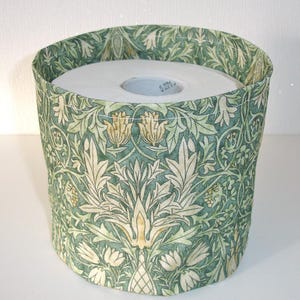 May include: A cylindrical toilet roll holder made of fabric, featuring a botanical pattern in green, cream, and yellow. The holder contains a roll of white toilet paper. The fabric design showcases detailed floral and leaf motifs.