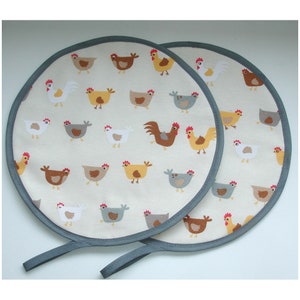 May include: Two round, grey-trimmed pot holders with a white background and a repeating pattern of colourful chickens. The pot holders have grey fabric loops for hanging.