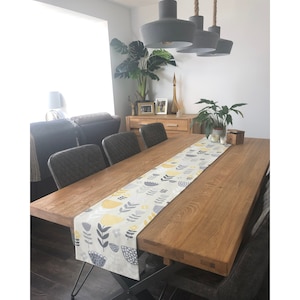 May include: A wooden dining table with a grey and yellow floral table runner. The table is set with chairs and has a light wood finish.