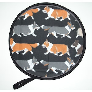 May include: Round black hot pad with a black handle. The hot pad is covered in a fabric with a repeating pattern of colourful corgi dogs running.