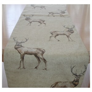 May include: A beige table runner with a repeating pattern of brown deer with antlers. The deer are facing to the right and are printed on a textured fabric.