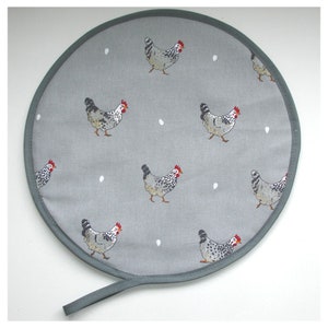 May include: A round grey and white fabric hot pad with a grey trim and a black handle. The hot pad features a repeating pattern of white chickens with black and grey markings and red combs. The background is a light grey with white dots.