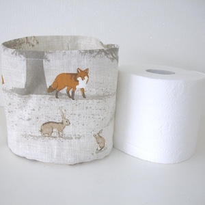May include: A cylindrical fabric storage basket featuring a light beige linen-like texture with a printed pattern of orange foxes and brown rabbits in a forest setting, positioned next to a standard white roll of toilet paper.