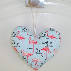 May include: A heart-shaped, light blue fabric ornament with a pink flamingo pattern. The ornament has a white string for hanging.