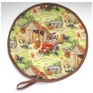 May include: Round brown and green fabric pot holder with a farm scene. The pot holder features a red tractor, cows, chickens, and a barn. The pot holder has a brown strap handle.