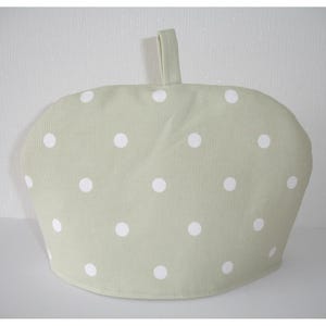 May include: A sage green tea cosy featuring a repeating pattern of white polka dots. The fabric cover has a rounded shape with a small fabric loop attached to the top centre for hanging or lifting, designed to insulate a teapot and retain heat.