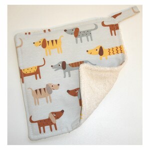 May include: A light grey kitchen towel with a white terry cloth backing. The towel features a repeating pattern of brown, yellow, and grey dogs with different coloured spots. The towel has a loop for hanging.