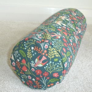 May include: A cylindrical pillow with a teal background, covered in a floral pattern with red flowers, green leaves, and birds. The pillow is approximately 12 inches long and 6 inches in diameter. The pillow is made of fabric.
