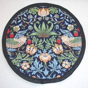 May include: Circular navy blue fabric trivet with a black trim. The design features birds and floral patterns in green, orange, and blue. Two birds are shown holding strawberries.