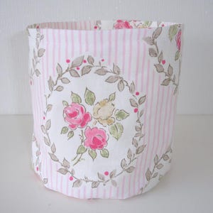 May include: A cylindrical fabric storage basket featuring a white background with vertical light pink stripes. The exterior is decorated with a repeating pattern of floral wreaths containing pink and yellow roses surrounded by green leaves and small red dots.