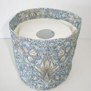 May include: A cylindrical fabric cover featuring a blue, beige, and white botanical pattern with intricate leaf and flower motifs. The cover is designed to fit around a standard white roll of toilet paper, which is visible inside the opening.