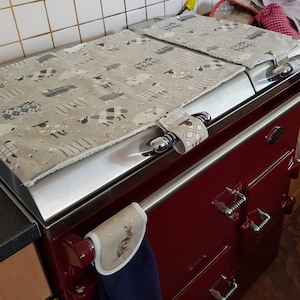 May include: A red AGA range cooker with two oven doors and a silver top. The top is covered with two grey and white patterned oven mitts with a sheep design. A blue and white tea towel with a rabbit design is hanging on the side of the cooker.