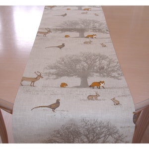 May include: A beige linen table runner featuring a woodland scene with a brown deer, a red fox, a grey squirrel, a brown rabbit, and a brown pheasant. The background features a grey tree with brown branches.