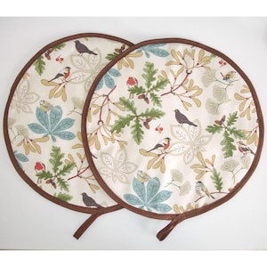 May include: Two round fabric pot holders featuring a cream background with a repeating pattern of various small birds, green oak leaves, light blue chestnut leaves, and tan branches. Each circular pot holder has a brown fabric trim and a small loop.