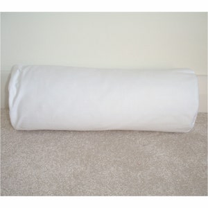 May include: A white cylindrical pillow, made of soft fabric, is lying on a light brown carpet.