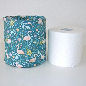 May include: A cylindrical fabric storage basket featuring a teal background with a repeating pattern of pink flamingos, green leaves, and small flowers, positioned next to a standard white roll of toilet paper for a size comparison.