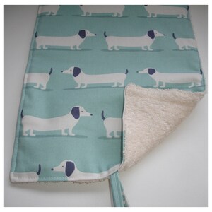 May include: A teal blue fabric placemat with a white and navy blue dachshund pattern. The placemat is reversible and has a soft, white terry cloth backing.