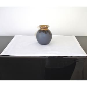 May include: A small, round, gray ceramic vase with a gold rim sits on a white linen table runner. The vase is on a black surface.