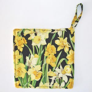 May include: Square pot holder with a black background and a pattern of yellow and white daffodils with green stems and leaves. The pot holder has a yellow loop for hanging.